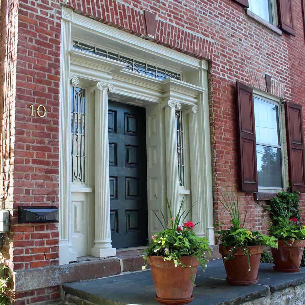This Greek revival entryway features ionic columns on either side of the door, along with leaded glass toplight and sidelights, and a keystone detail above the door and each window in the building's brick facade. The door itself is heavy wood, and the small area outside is lined with slate and dotted with container plantings.